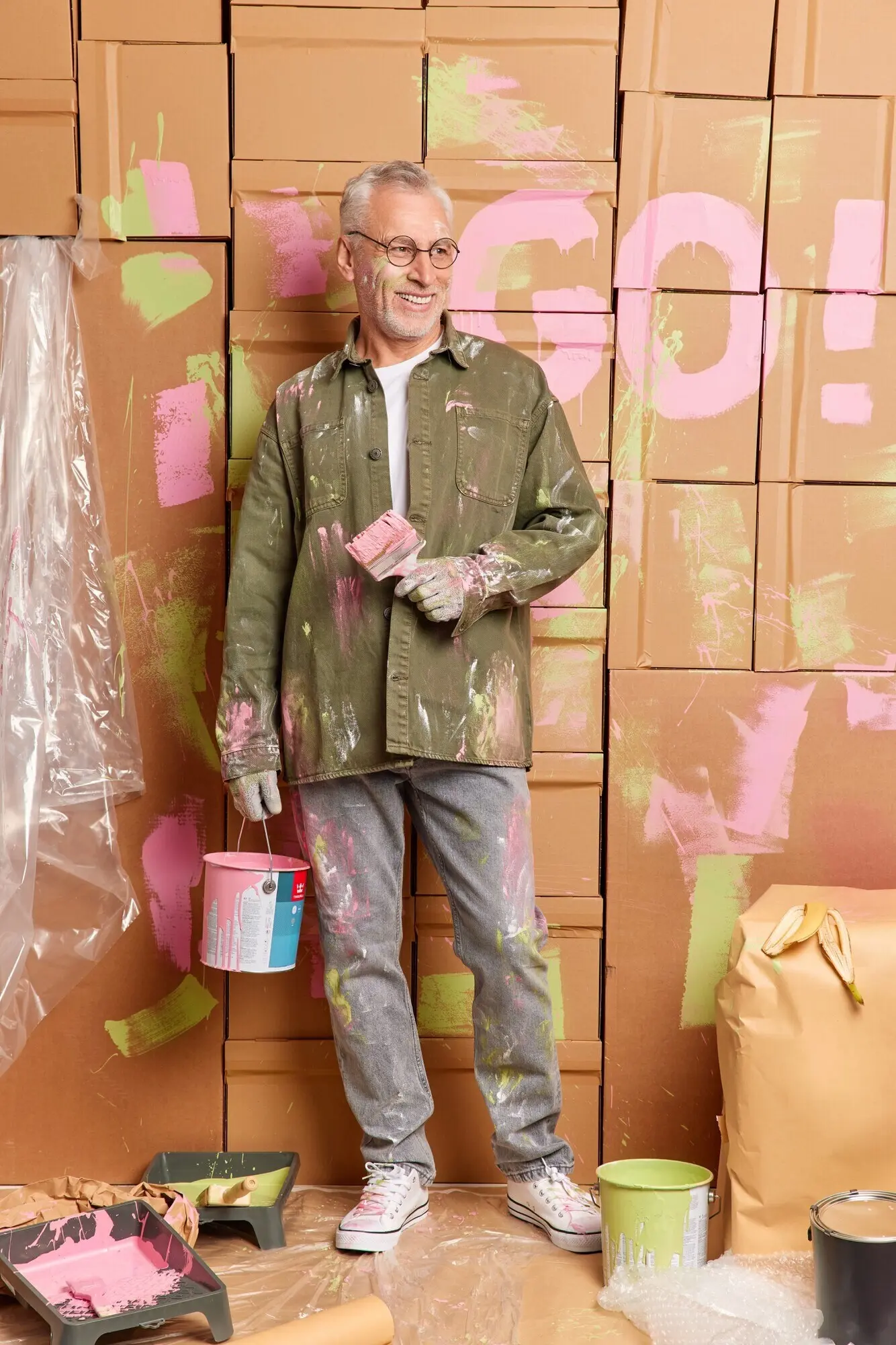 Fröhlicher älterer Mann hält einen Farbeimer und einen Pinsel, führt Reparaturen durch, blickt gut gelaunt zur Seite, trägt legere Kleidung und ist zu Hause mit Renovierungsarbeiten beschäftigt. Konzept: Umbau, Hausverbesserung, Instandhaltung.